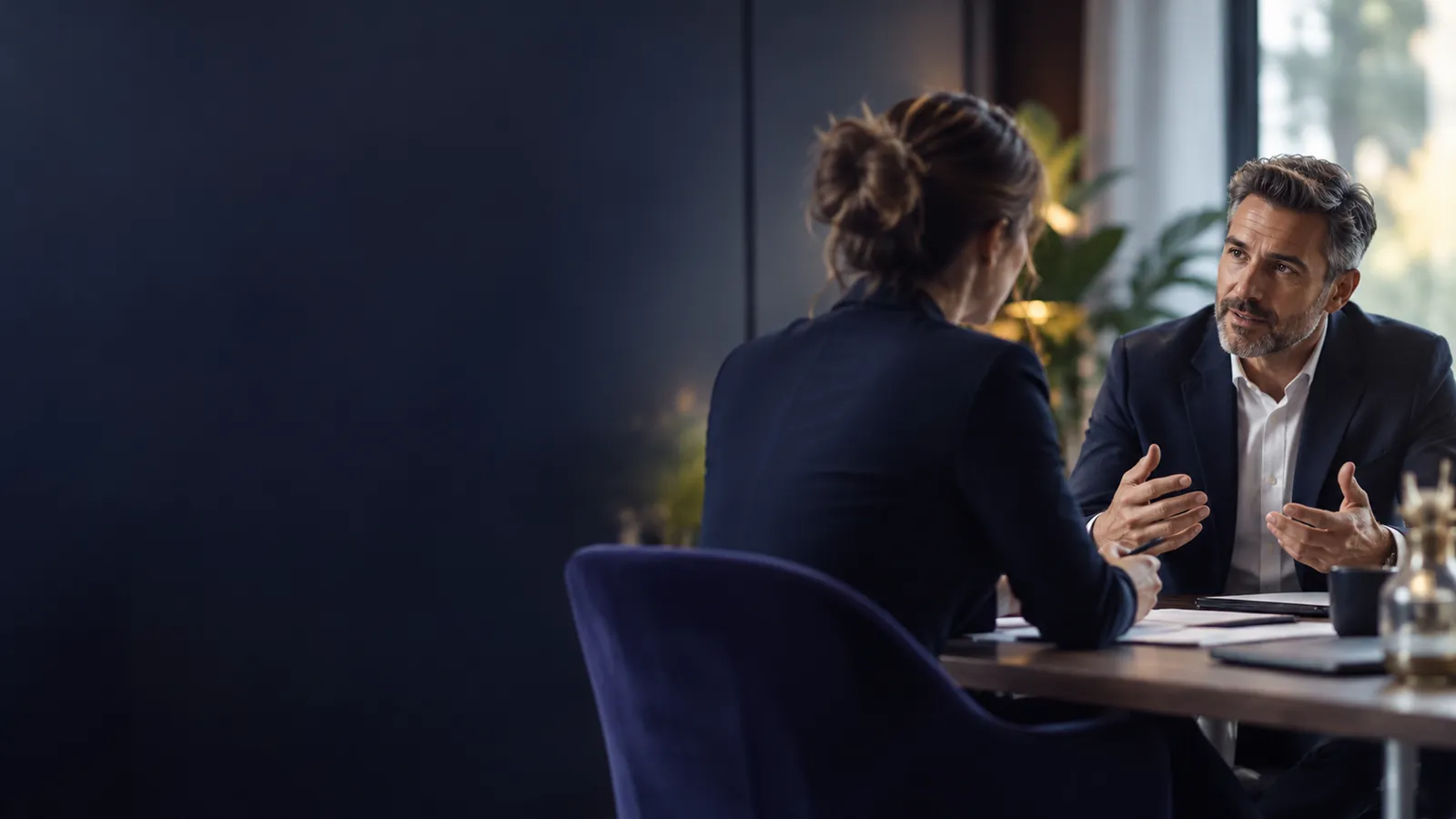 Entretien entre une consultante vue de dos et un dirigeant attentif autour d'une table, dans un bureau aux tons sombres