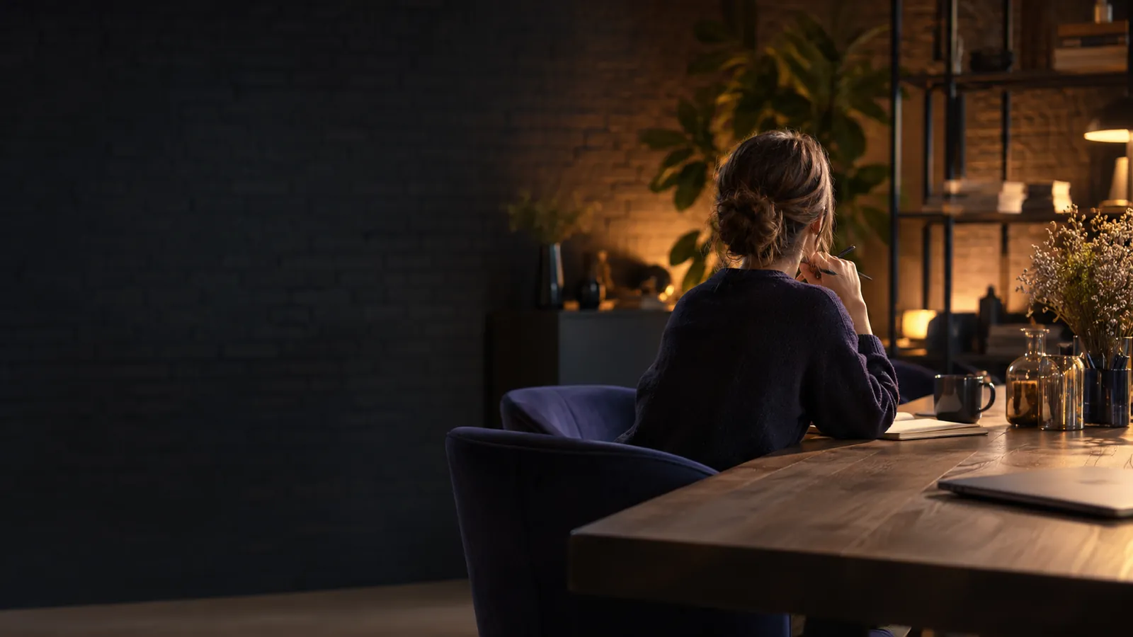 Personne travaillant à une grande table en bois dans un bureau aux murs de brique sombre, lumière de fin de journée