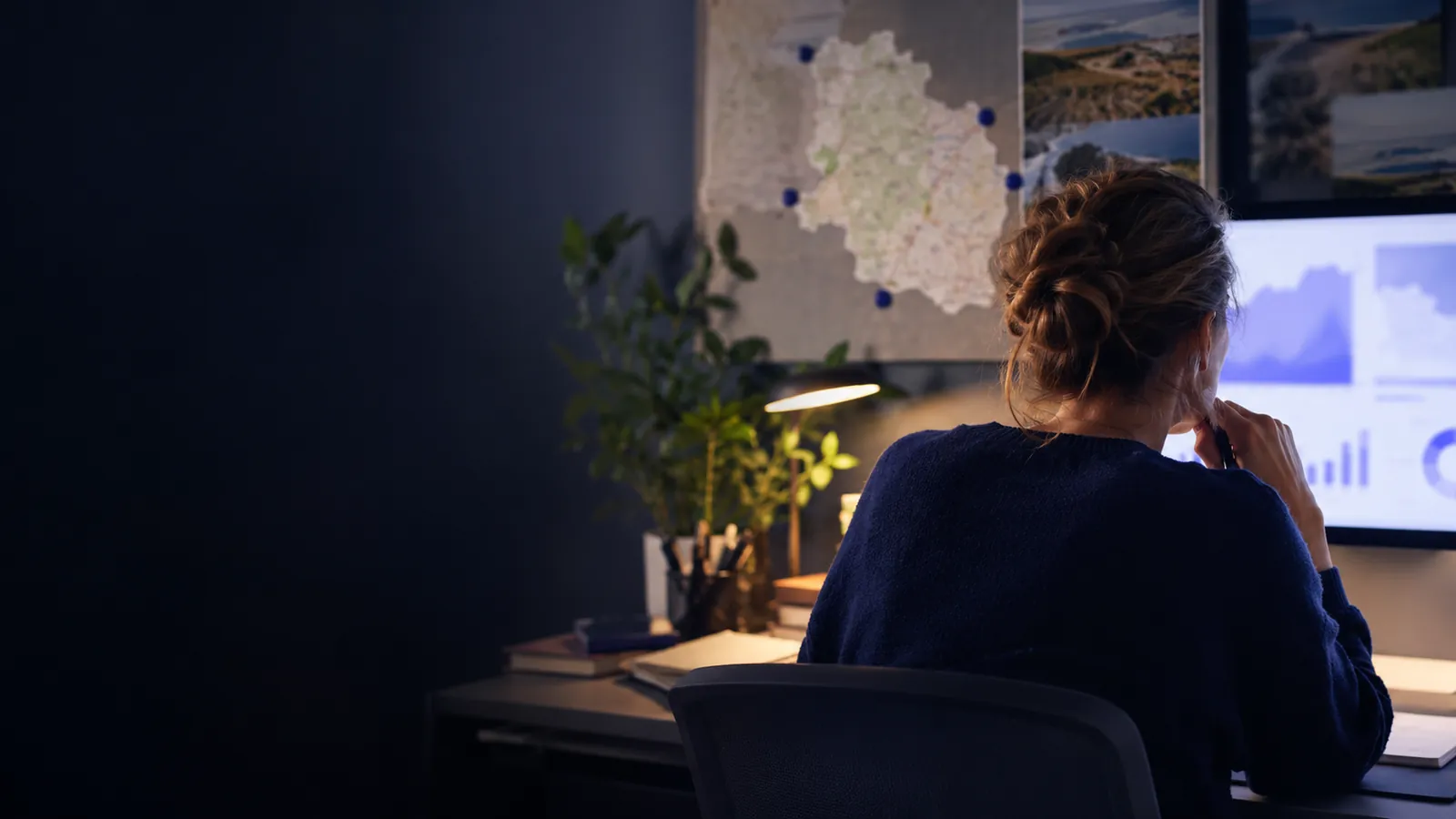 Femme vue de dos devant un écran de graphiques, carte régionale punaisée au mur de son bureau en soirée