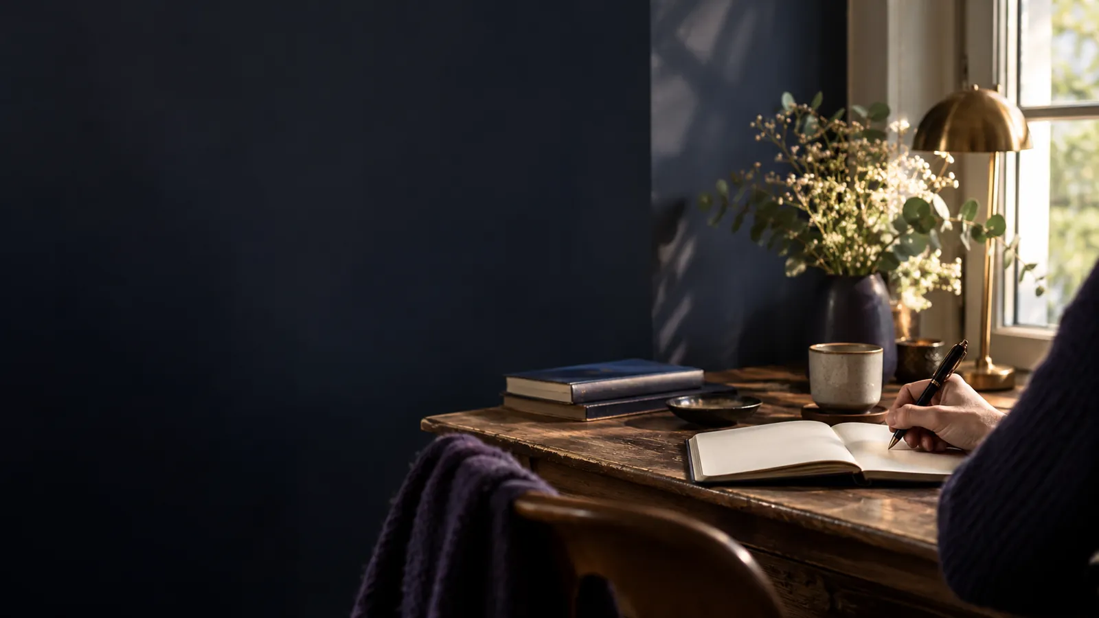 Personne écrivant dans un carnet à un bureau près d'une fenêtre, bouquet de fleurs et mur sombre