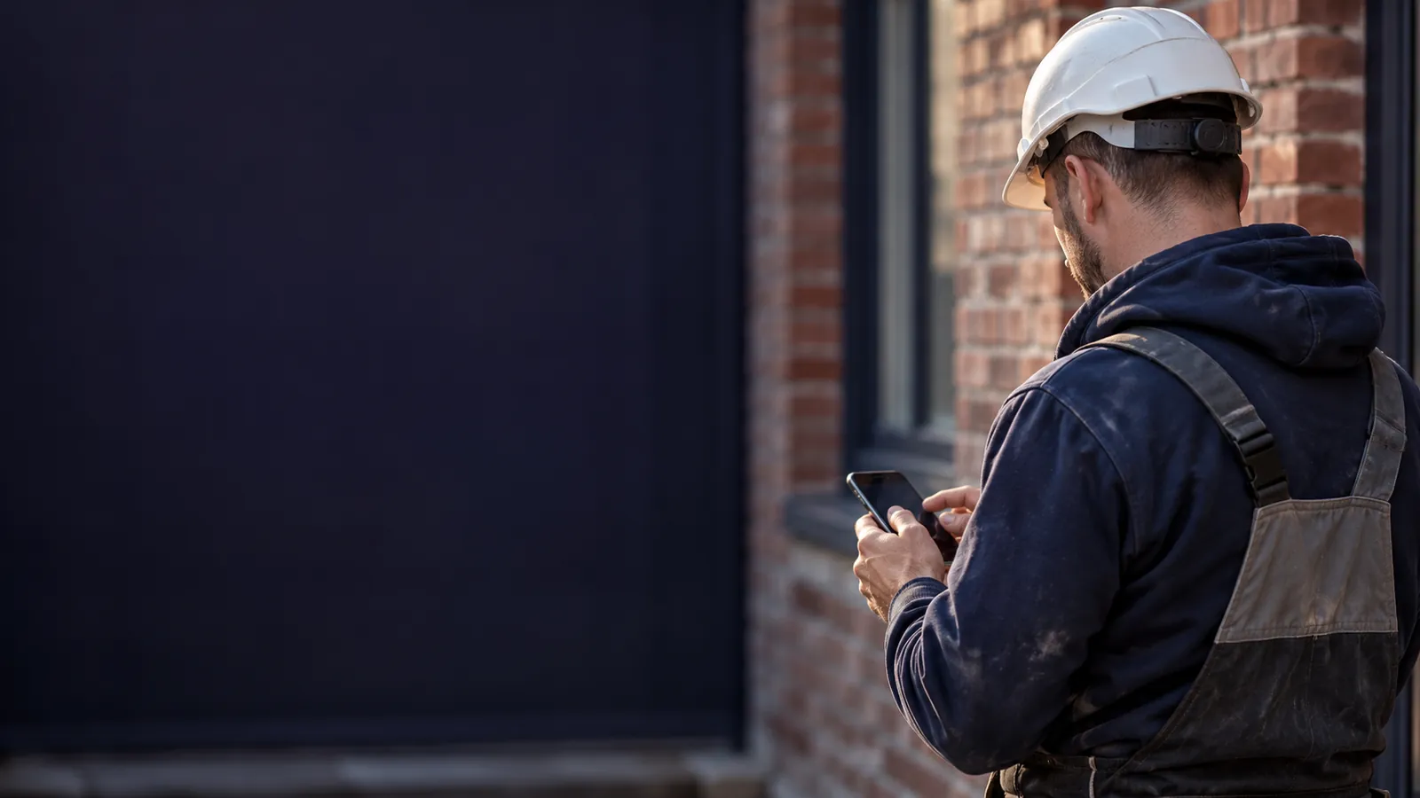 Artisan en tenue de chantier consultant son téléphone devant une façade en briques typique du nord de la France