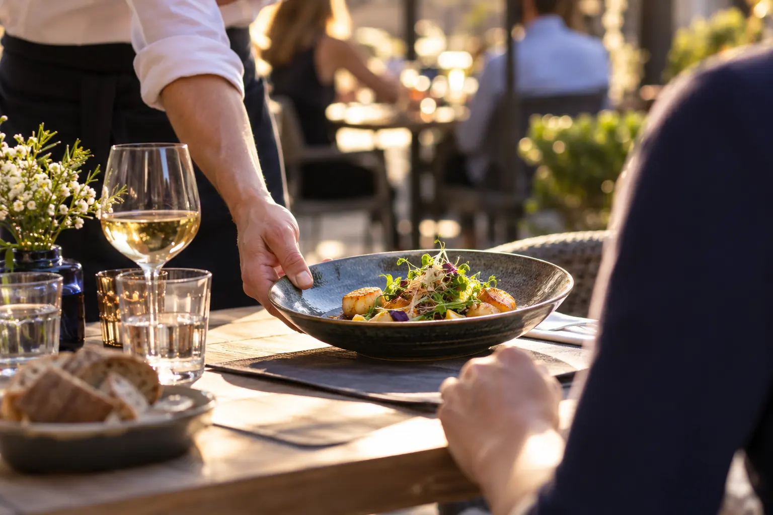 Serveur déposant une assiette garnie sur une table en terrasse, avec verre de vin blanc et pain, au soleil couchant